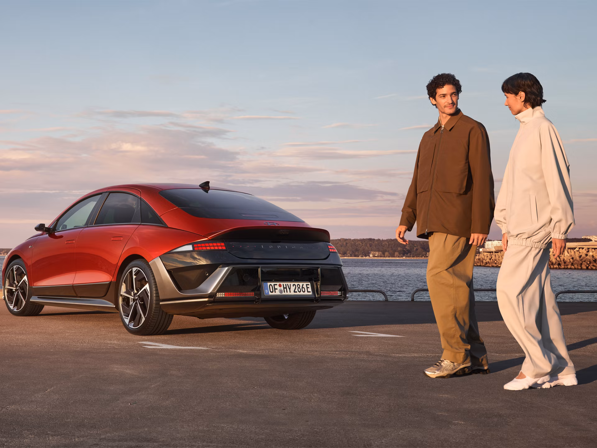 Muškarac i žena prolaze pored crvenog automobila Hyundai IONIQ 6 N Line parkiranog uz obalu.