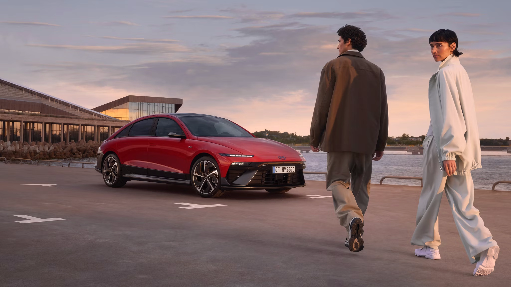 Muškarac i žena prolaze pored crvenog automobila Hyundai IONIQ 6 N Line parkiranog uz obalu.